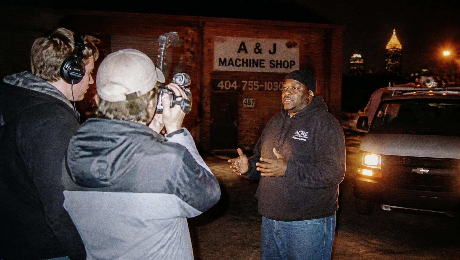 A two-person crew filming a machine shop owner outside his shop at night in Atlanta